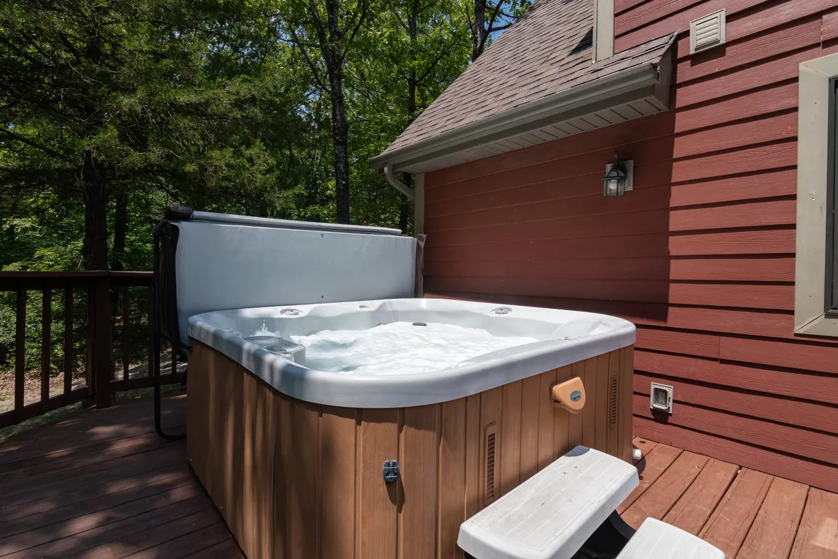 A serene view of the deck area with a prominent hot tub and natural wooden elements, set against a backdrop of tall trees. The setting emphasizes the peaceful isolation and immersion in the surrounding forest.