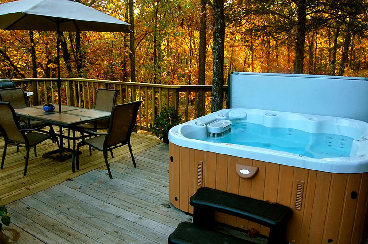 Featuring a private hot tub nestled on a wooden deck surrounded by trees, this tranquil outdoor space invites relaxation in nature. The adjoining dining table suggests al fresco meals or friendly gatherings, all within a peaceful, wooded setting.