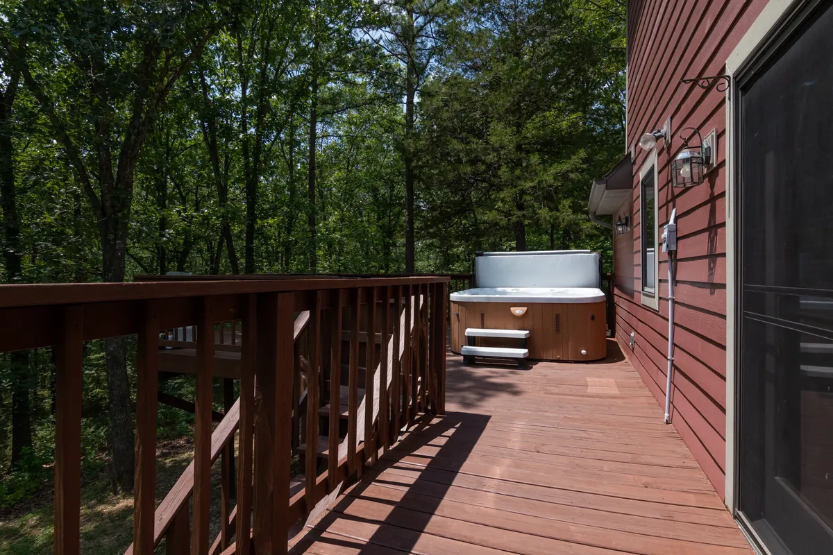 An exterior shot of a wooden deck featuring a bubbling hot tub, surrounded by trees. The space is designed for relaxation and recharging, with ample room for enjoying the outdoors in privacy.