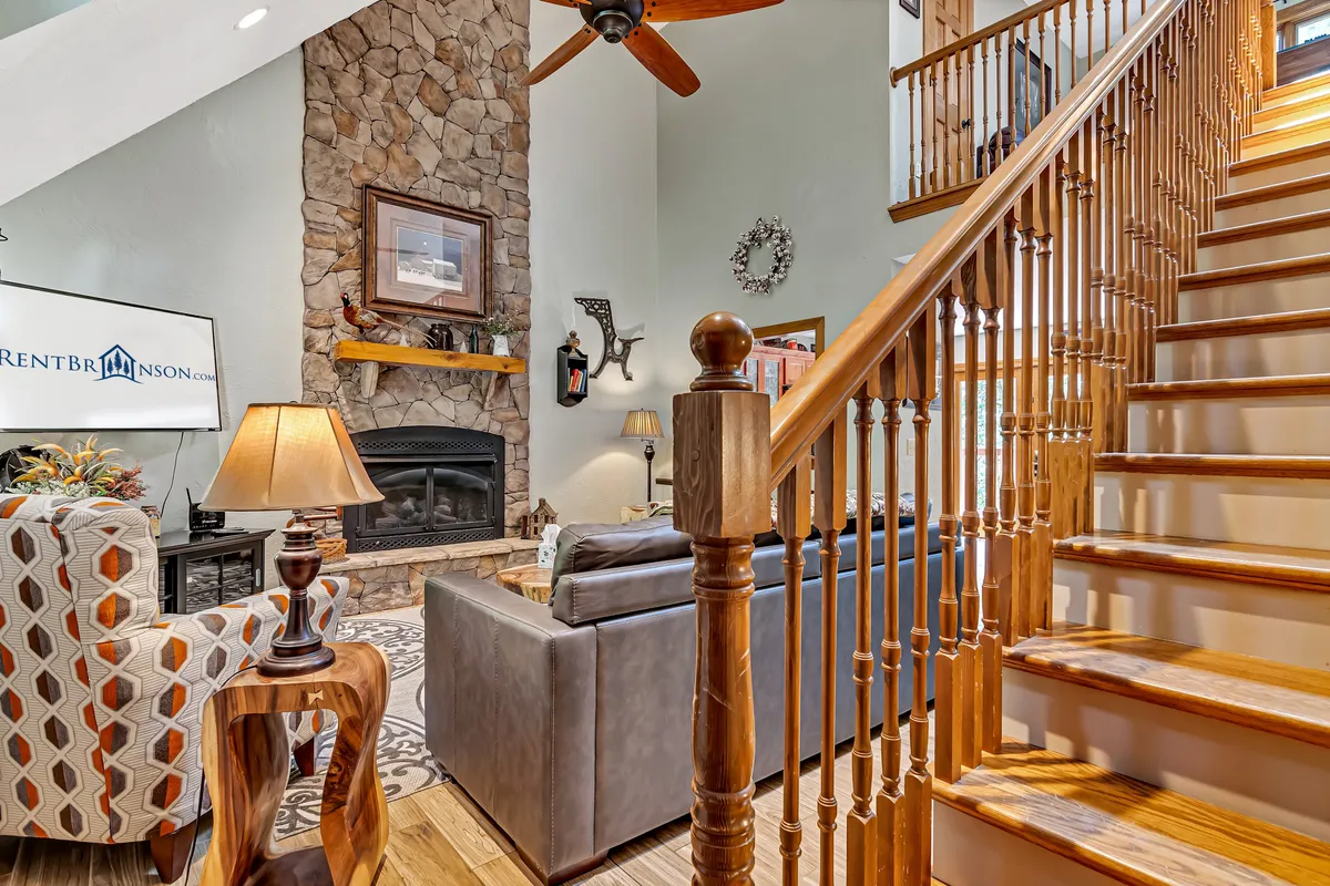 A welcoming living area with a prominent stone fireplace, wooden staircase, plush seating, and tasteful wall art. Natural light streams in, creating a warm and inviting atmosphere ideal for gathering and relaxation. The scene exudes both comfort and style, highlighting the fireplace as the central gathering point.