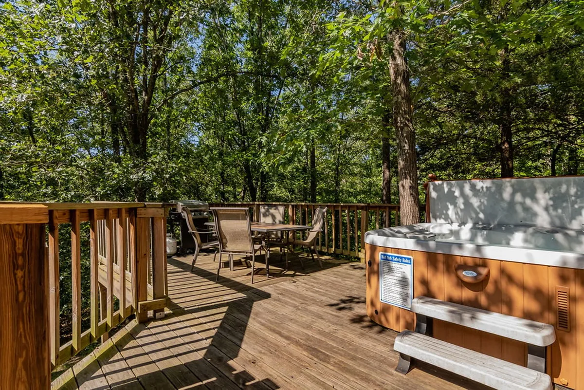 A wide, inviting deck scene featuring a hot tub, a dining area with a table and chairs, and views of the lush woods beyond. The setup is perfect for socializing, relaxing, or enjoying meals surrounded by nature.