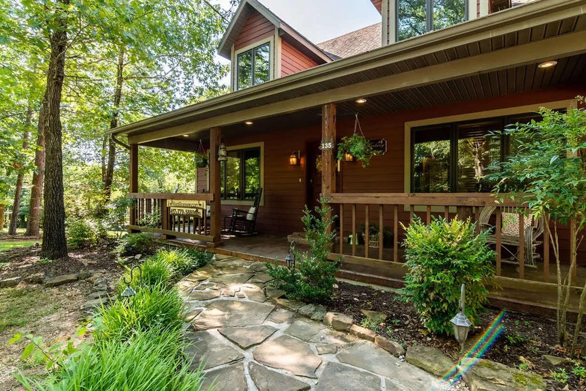 A welcoming front porch with rocking chairs and a stone pathway is framed by lush greenery, suggesting a perfect spot for enjoying sunsets, morning coffees, or casual gatherings. The setting is calming and speaks to both relaxation and social activity within a natural environment.