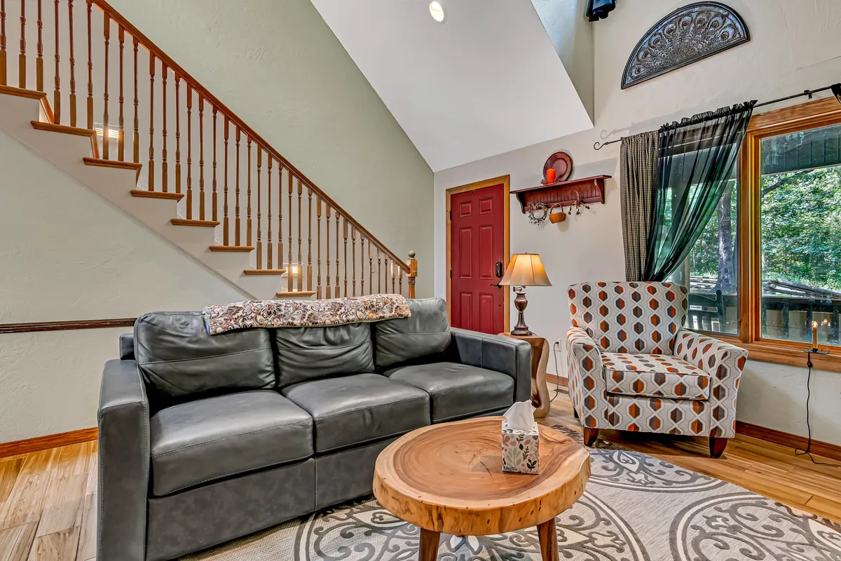 A cozy living space with a plush sofa, wooden coffee table, armchair, and staircase leading to a loft. Large windows fill the room with sunlight, creating a bright and welcoming environment ideal for morning coffee, reading, or socializing. The window view connects the interior to the surrounding woods, echoing the 'sun-kissed' and 'expansive views' described in the section.