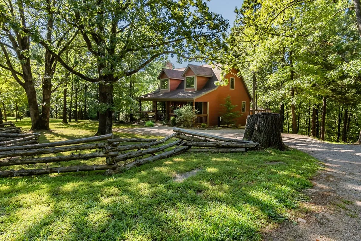 The hero image showcases a charming orange cottage nestled amid lush trees, embraced by a rustic wooden fence. The peaceful natural setting hints at expansive grounds perfect for exploring, while the porch invites relaxation and sunset views.