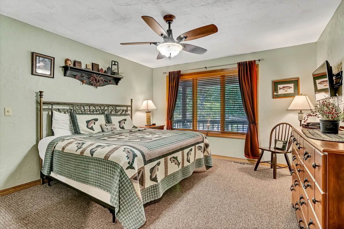 A tranquil bedroom with a patterned quilt on the bed, a wooden dresser, and a window with curtains letting in natural light. The decor features warm wood tones and a ceiling fan above, creating a cozy atmosphere ideal for rest and relaxation.