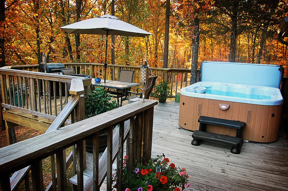 A tranquil outdoor scene featuring a wooden deck nestled among trees, with a bubbling hot tub, an outdoor dining set, a propane grill, and colorful flower pots. The deck is surrounded by lush greenery, offering immersive woodland views and a serene atmosphere.