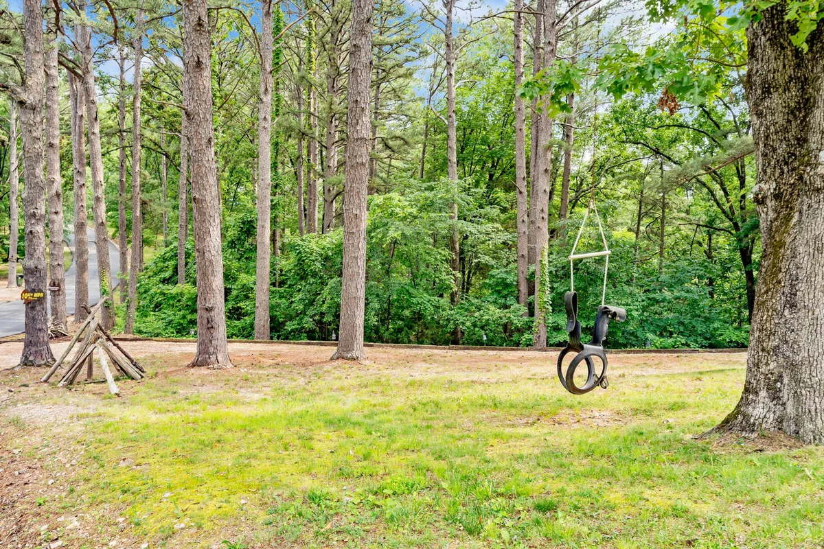 A cleared grassy area under tall trees is centered around a playground swing set, providing a playful and safe environment for children. The natural setting emphasizes opportunities for outdoor recreation and exploration.