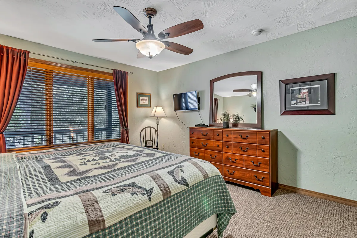 A cozy bedroom featuring a bed with a patterned quilt, a large window offering nature views, and a wooden dresser. The room is filled with soft, natural light and decorated in soothing tones, evoking comfort and privacy.