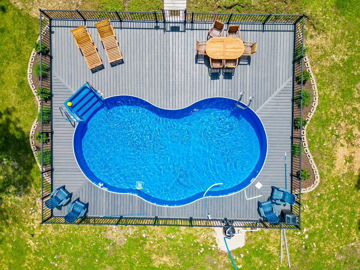 This image showcases a sparkling swimming pool framed by a wooden deck, outdoor dining table, and lounge chairs, all set against a background of lush trees. The scene exudes a welcoming, resort-style atmosphere ideal for community gatherings, sunbathing, and outdoor fun.